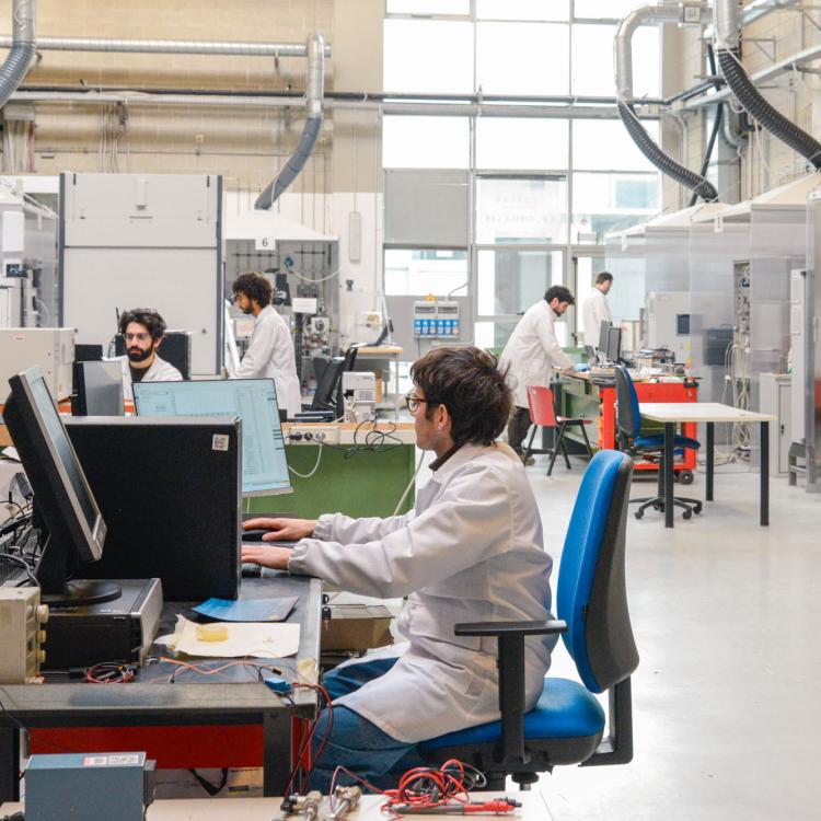 Researchers in lab coats working at workstations and equipment inside a large modern scientific laboratory.